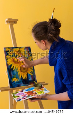 Girl artist paints sunflowers oil paints on canvas. She is wearing blue sweater. Woman is holding brush and palette with paints. She rubs paint with her finger.