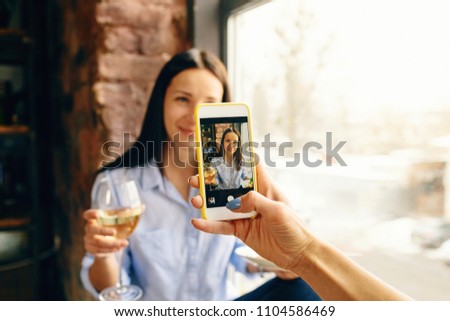 Woman taking photo her beautiful young girl friend with glass of wine on smartphone