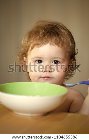 Kid play. Portrait of cute sweet little boy with blonde curly hair and round cheecks eating from green plate with spoon closeup, vertical picture