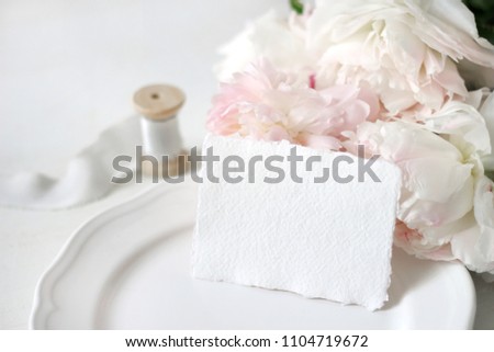 Bright wedding or birthday stationery mockup scene with a handmade paper greeting card, porcelain plate, spool of silk ribbon and pink peony flowers a white table background. Feminine styled stock pho