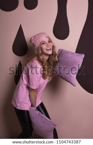 Adorable happy blonde woman playing with pillows on a pink studio background