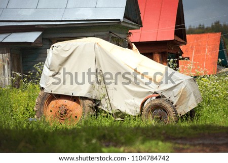 The old tractor in the cover is in the summer day on the background of village houses.