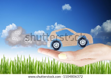 CO2 pollution concept with 
cardboard model  car with exhaust smoke clouds in woman hand, natural background with green grass and clear blue sky 
