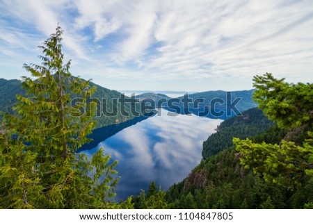 Horizontal photo of a View of Lake Crescent in Olympic National Park from Storm King with blue skies and clouds reflecting in the blue water. Mountains and hills in the background with evergreen trees