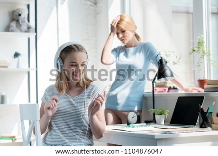 happy teen daughter listening music while her disappointed mother standing on background