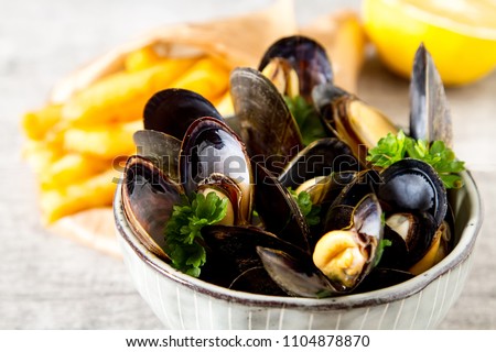 Mussels with herbs in a bowl with lemon and French fries on a white wooden board. Seafood. Food at the shore of the French Sea. Dark background Royalty-Free Stock Photo #1104878870