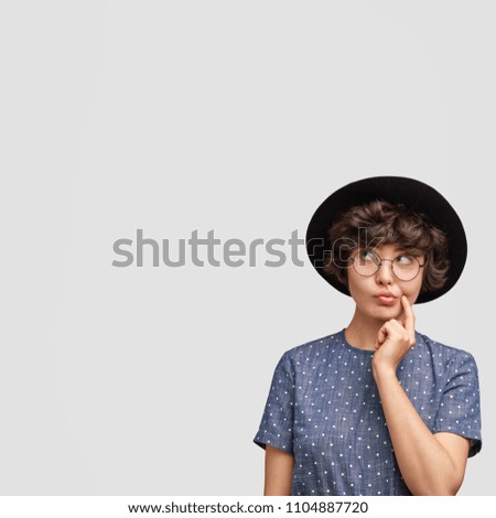 Portrait of attractive brunette female loos thoughtfully upwards, wears elegant black hat, keeps finger near mouth, stands against white background with blank space for your promotional text