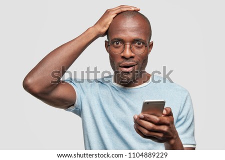 Attractive dark skinned male with stubble, holds smart phone, stunned to recieve text message, connected to wireless internet, surfes web page, isolated on white background. Diversity and technology