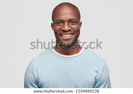 Middle aged cheerful dark skinned male with shining smile, wears light blue sweater, round spectacles, achieves everything by himself, stands against white studio wall, has relaxed face expression Royalty-Free Stock Photo #1104889238