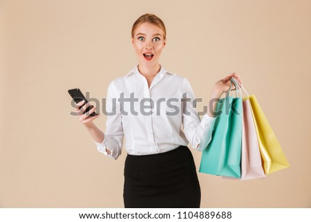 Photo of emotional young business woman standing isolated over beige wall backgound looking camera holding shopping bags using mobile phone.