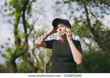 Young athletic tired beautiful brunette girl in black uniform and cap talking on mobile phone during training, keeping hand near head in city park outdoors. Fitness, healthy lifestyle concept