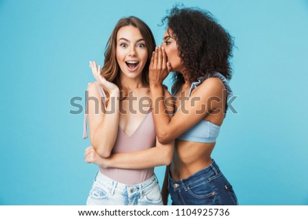 Two cheerful young girls dressed in summer clothes whispering secrets isolated over blue background Royalty-Free Stock Photo #1104925736