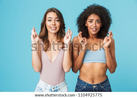 Two excited young girls dressed in summer clothes holding fingers crossed for good luck isolated over blue background