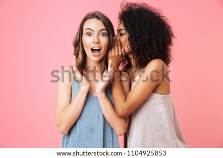 Two excited young girls dressed in summer clothes whispering secrets isolated over pink background Royalty-Free Stock Photo #1104925853