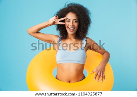 Portrait of a happy african girl dressed in swimsuit holding infatable ring and showing peace gesture isolated over blue background