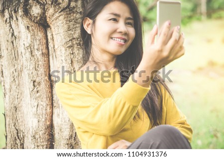 attractive Asian woman sitting under the tree in a park with smart phone in hand taking picture or selfie and smile at a camera 