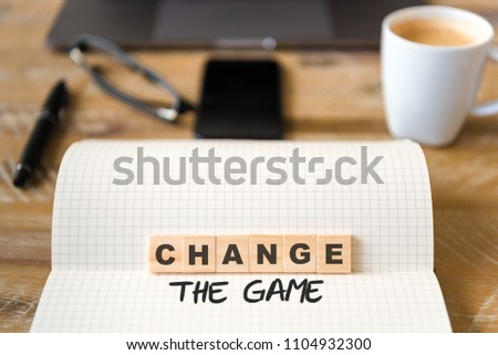 Closeup on notebook over wood table background, focus on wooden blocks with letters making Change the Game text. Concept image. Laptop, glasses, pen and mobile phone in defocused background