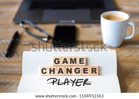 Closeup on notebook over wood table background, focus on wooden blocks with letters making Game Changer not Player text. Concept image. Laptop, glasses, pen and mobile phone in defocused background