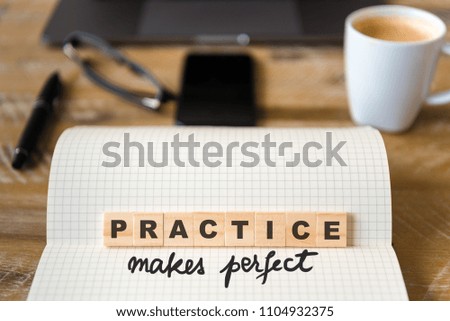 Closeup on notebook over wood table background, focus on wooden blocks with letters making Practice Makes Perfect text. Concept image. Laptop, glasses, pen and mobile phone in defocused background