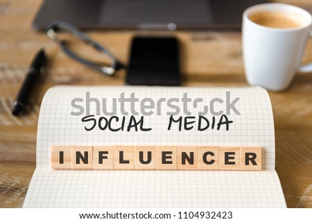 Closeup on notebook over wood table background, focus on wooden blocks with letters making Social Media Influencer text. Concept image. Laptop, glasses, pen and mobile phone in defocused background