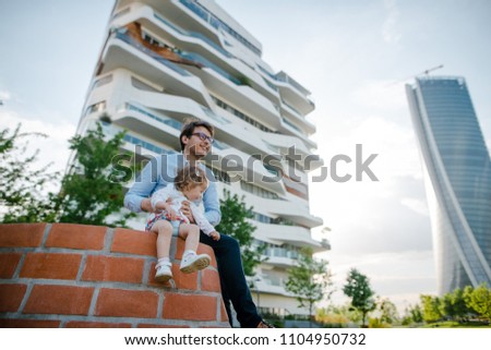 Young father and his daughter sitting on the brick sculpture in the city park near the modern skycrapers. Sunny summer day. Happy family