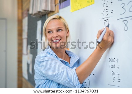 Teacher teaching how to count on whiteboard in classroom. Smiling blonde woman explaining additions in column in class. Math’s teacher explaining arithmetic sums to elementary children. Royalty-Free Stock Photo #1104967031