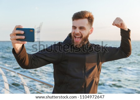 Excited young sportsman taking selfie with mobile phone while standing at the beach and flexing biceps
