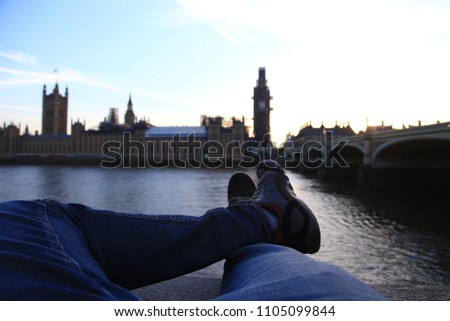 POV image of legs as the photographer sits on the edge of the River Thames in London England looking at a silhouette of the English Parliament in Westminster at sunset.