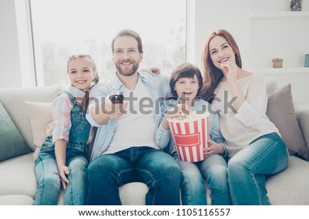 Portrait of mama daddy two kids looking tv sitting on sofa having pop-corn in hands using remote-control searching interesting cartoons enjoying cinema indoor. Weekend holiday free time concept