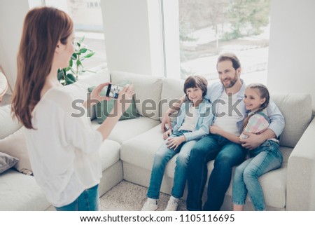 Portrait of wife shooting photo for memory on smart phone's quality camera of husband and two kids sitting on couch embracing wearing casual jeans outfits enjoying time together