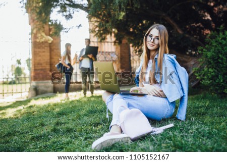 A very pretty girl student looking to the camera sitting in a campus and studying on a laptop. In the background, a group of people was blurred.