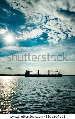 Freight Ship silhouetted with a satburst sun behind it against a cludy blue sky and dark blue water
