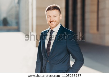 Portrait of successful intelligent businessman wears formal black suit, has positive expression, poses outdoor, rejoices successfully signed contract with business partners. Cheerful entrepreneur