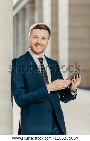 People, work and technology concept. Handsome male economist searches information online on digital tablet, prepares for meeting with staff, works on financial report, dressed in formal clothing