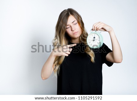 beautiful blond girl points finger at retro alarm clock, studio photo on background