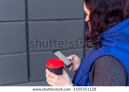 close-up of hipster girl holding coffee and smartphone with blank screen for your logo or text message, girl walking around town at night and drinking cappuccino