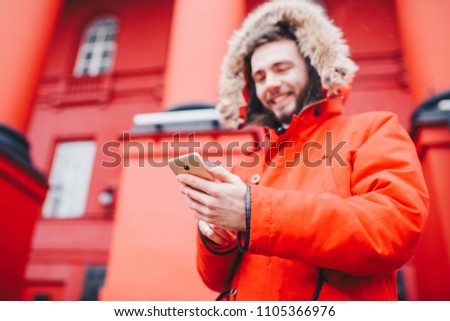 handsome young male student with toothy smile and beard stands on red wall background, facade of educational institution in red winter jacket with hood with fur, Uses finger on screen of mobile phone.