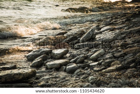 A rocky shoreline on the banks of Lake Ontario shimmers in the evening twilight. Small cresting waves flow toward large wet stones at water’s edge. Warm shades of gray, yellow and pale blue.