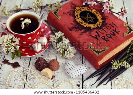 Red cup of flower tea with black candles, candies and witch book on table. Occult, esoteric and divination still life. Halloween background with vintage objects 