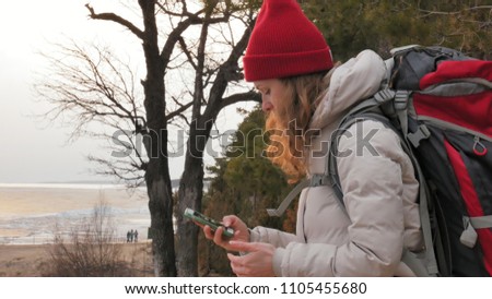 An attractive young woman in a red hat walks through the forest in early spring with a large tourist backpack and takes pictures on the phone 4k.