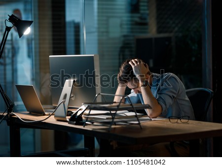 Young man in shirt working alone in office late sitting in lamplight at table and looking sleepy.  Royalty-Free Stock Photo #1105481042