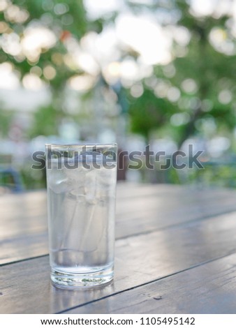 Closeup of a glass of water with ice, ready to be sipped, on an out door dinning table with green trees bokeh in the background
