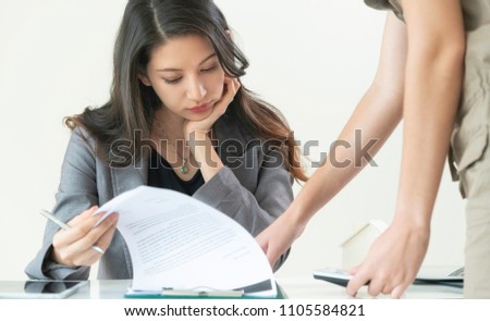 Businesswoman reads paper documents while discussing with another businesswoman standing in front of her partner at the office.