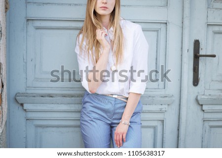 Thin young woman with long straight blonde hair wearing white shirt and blue trousers. Business outfit. Casual look. Standing next to big old blue door. Part of body look.