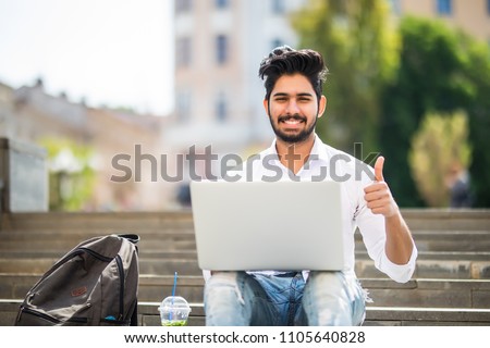 Happy indian student sitting on the stairs showing thumb up working on laptop, in the university campus. Royalty-Free Stock Photo #1105640828