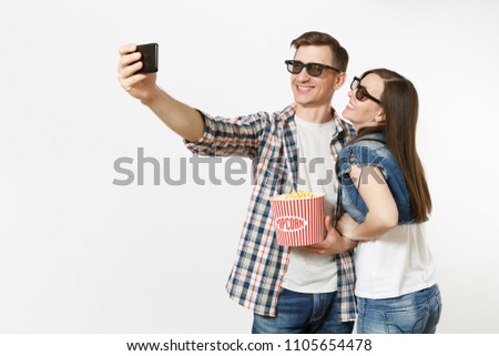Young couple, woman and man in 3d glasses watching movie film on date, holding bucket of popcorn and cup of soda or cola, doing selfie on mobile phone isolated on white background. Emotions in cinema