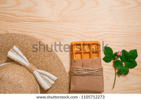 Minimal styled on wood background. Feminine desk top view with summer accessories: thatch hat, croissant waffle in a kraft package, rose flowers, flat lay