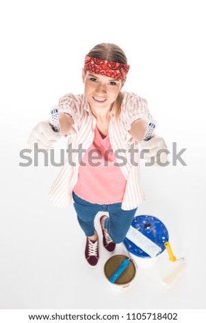 high angle view of young woman doing thumbs up gesture and standing near tin of paint,  paint roller and paint brush isolated on white background 