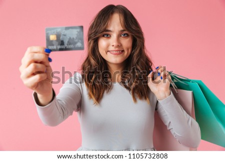 Photo of cute young pretty woman shopaholic standing isolated over pink wall background looking camera holding shopping bags and credit card.