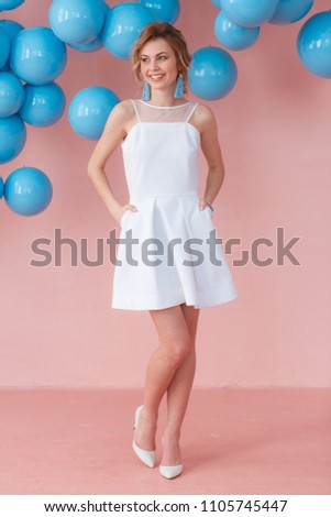 Young woman in white cocktail dress posing on pink wall background with blue bubbles hanging. Dreaming concept.
